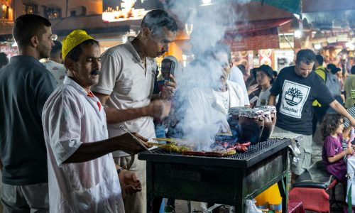 A man grills food on a smoky street food stall while others stand nearby in a busy outdoor market at night.