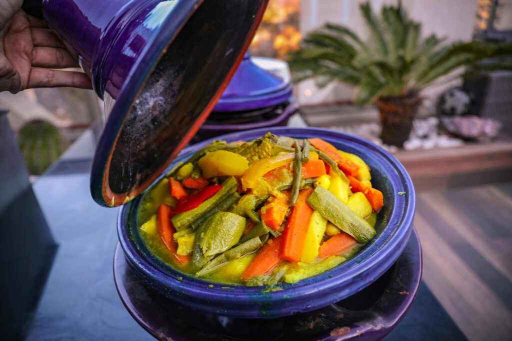 A hand lifts the lid off a purple tagine pot filled with cooked vegetables, including carrots, zucchini, and potatoes.