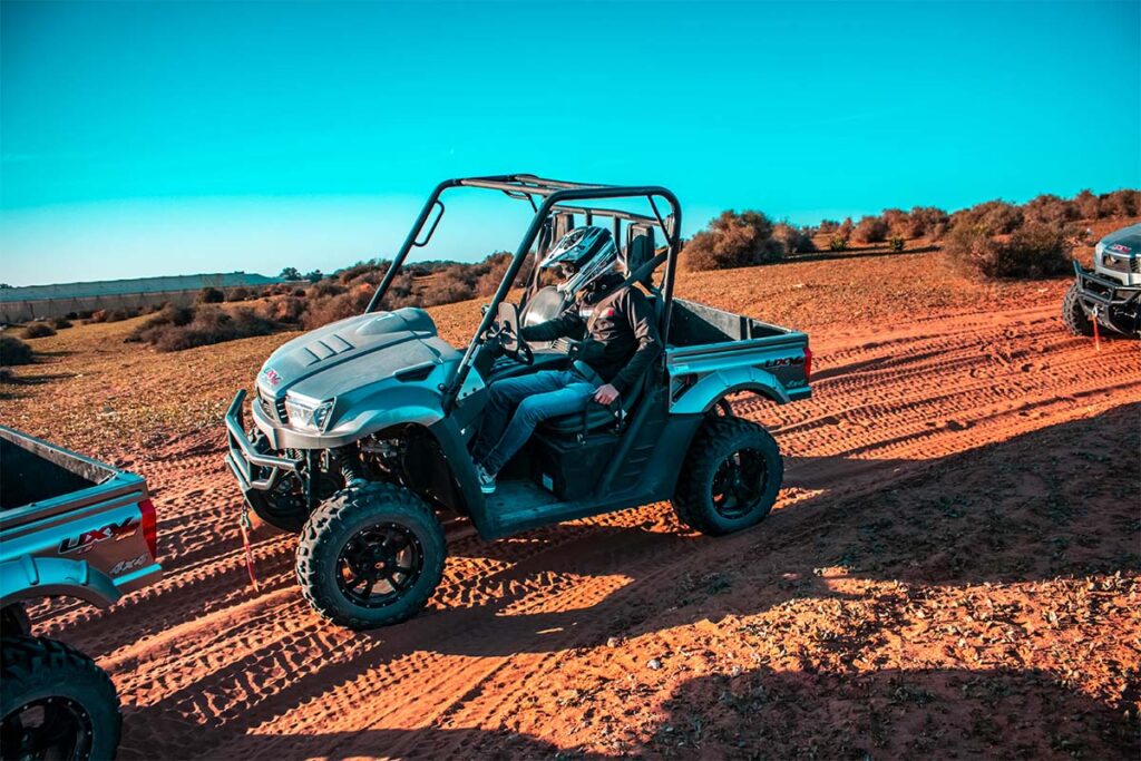 A person wearing a helmet drives an off-road utility vehicle on a dirt path surrounded by shrubs under a clear blue sky.