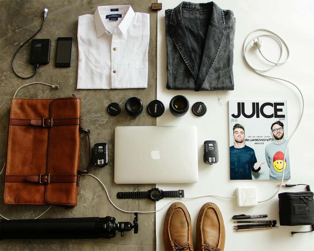 Neatly arranged items including a shirt, jacket, camera lenses, laptop, magazine, leather bag, shoes, tripod, watch, phone, chargers, and cables on a two-tone surface.