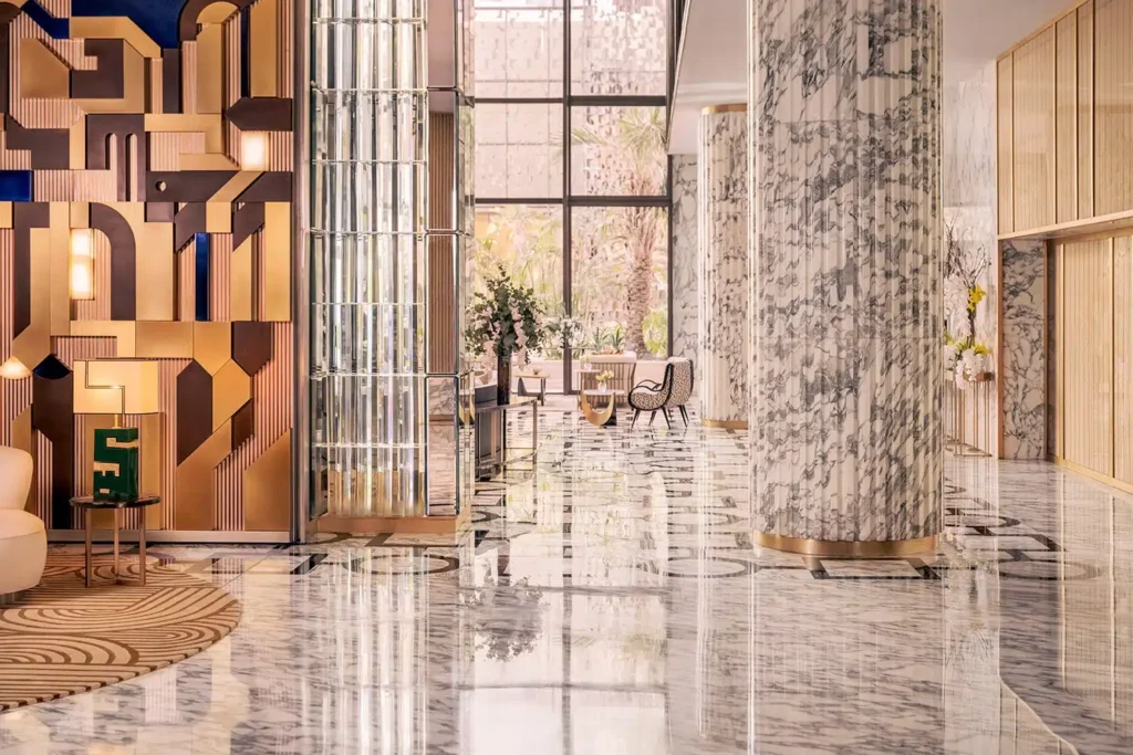 A spacious hotel lobby with marble floors, patterned columns, modern wall art, large windows, and stylish seating areas.