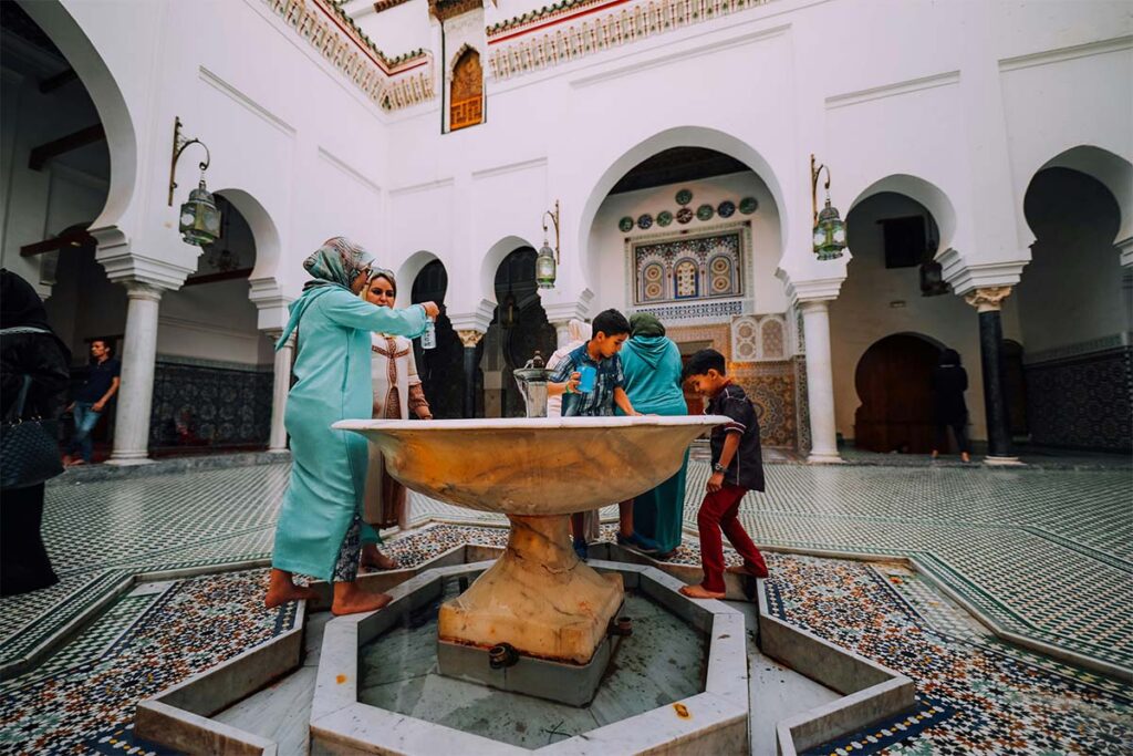 People in traditional clothing gather around a marble fountain in a tiled courtyard with arched architecture and decorative mosaic patterns.