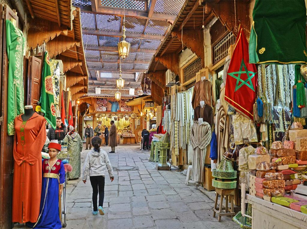 A narrow, covered market street lined with colorful fabrics, clothing, and Moroccan flags; people walk and shop along the cobblestone path.