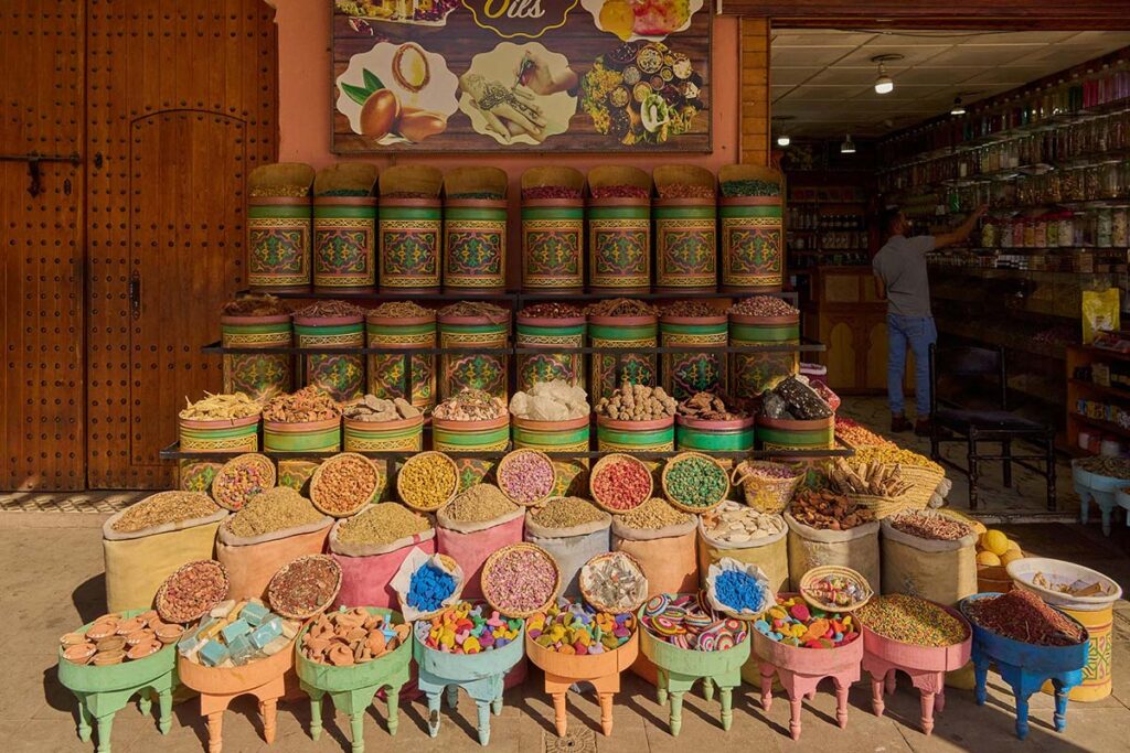 Colorful baskets and jars filled with various spices, herbs, and dried goods displayed outside a market shop in sunlight.