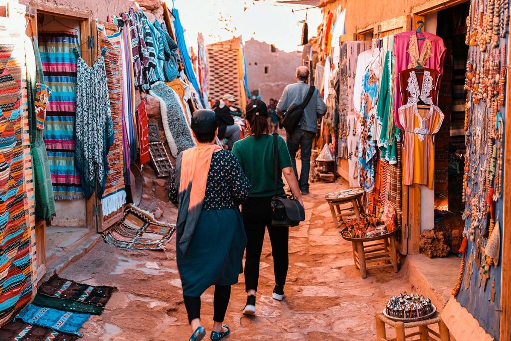 Two people walk through a narrow market street lined with colorful textiles, rugs, and handicrafts displayed on walls and stalls.