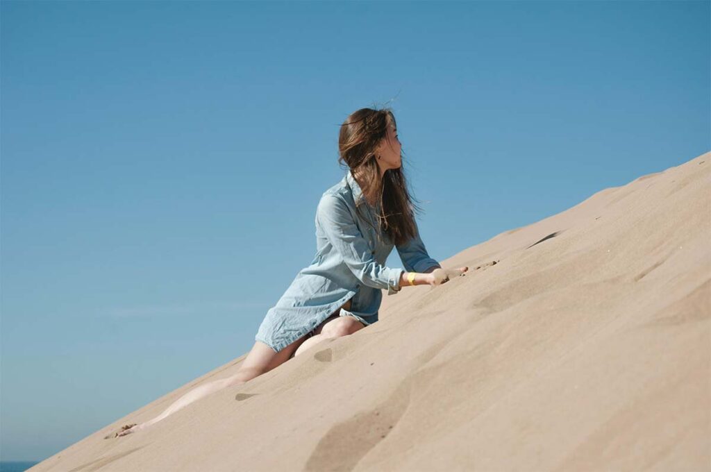 A woman in a blue dress climbs up a sandy dune under a clear blue sky.