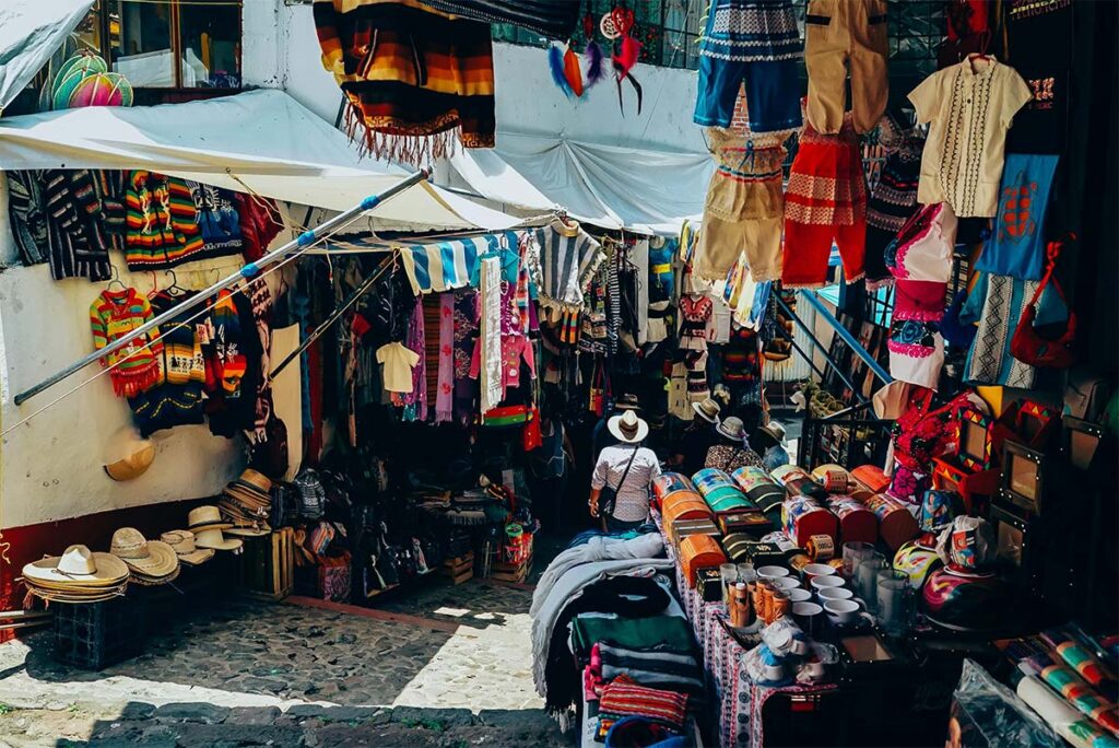 Outdoor market with colorful textiles, clothing, hats, and pottery on display. A person wearing a hat stands among the stalls on a cobblestone path.