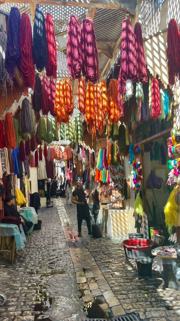 A narrow cobblestone alley lined with dyed yarns, as seen on a tour led by Siham Lahmine, founder of Morocco Travel Organizer, with people walking and working under dappled sunlight.