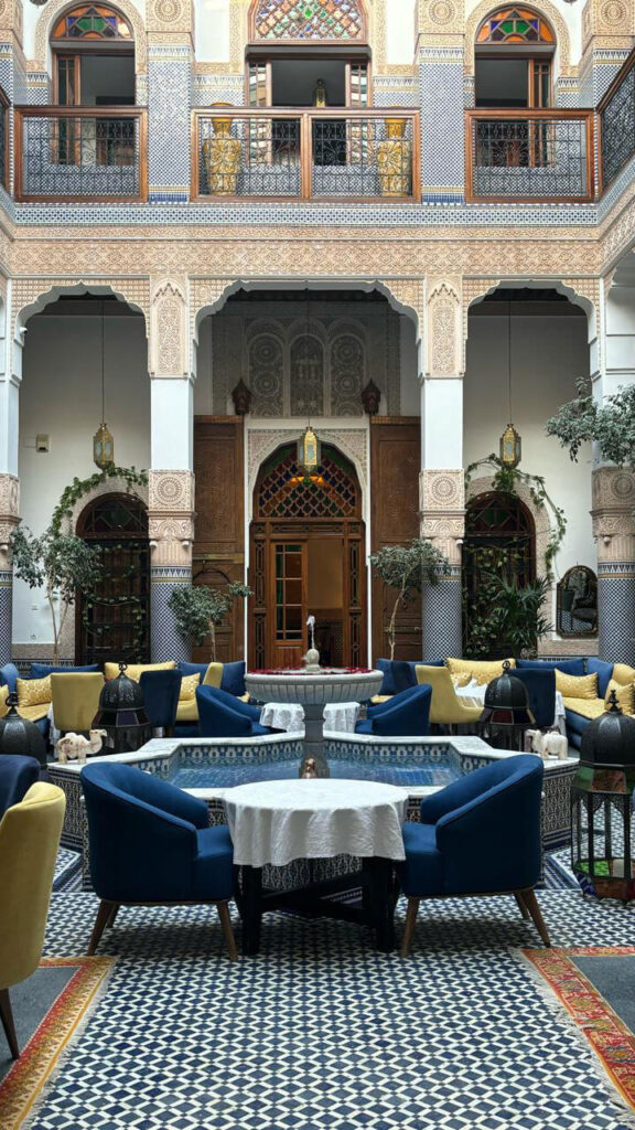 A courtyard with patterned tile flooring, a central round fountain, blue and yellow seating, ornate arches, and wooden balconies—perfect inspiration as you plan your luxury culinary or heritage trip to Morocco.