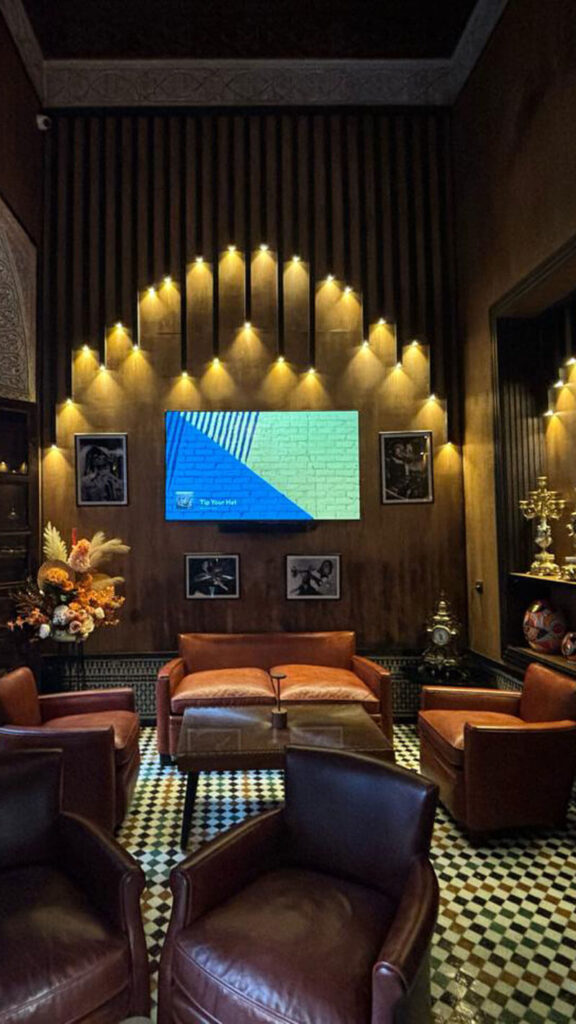 A lounge with brown leather chairs, a checkered tile floor, a large wall-mounted TV, framed photos, decorative vases, and warm accent lighting designed by Siham Lahmine, founder of Morocco Travel Organizer.