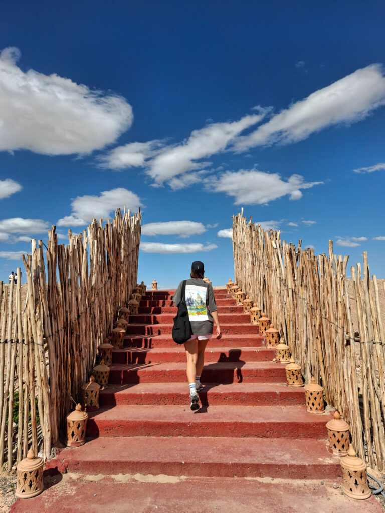 A person with a backpack walks up red steps lined with bamboo railings under a blue sky with scattered clouds.