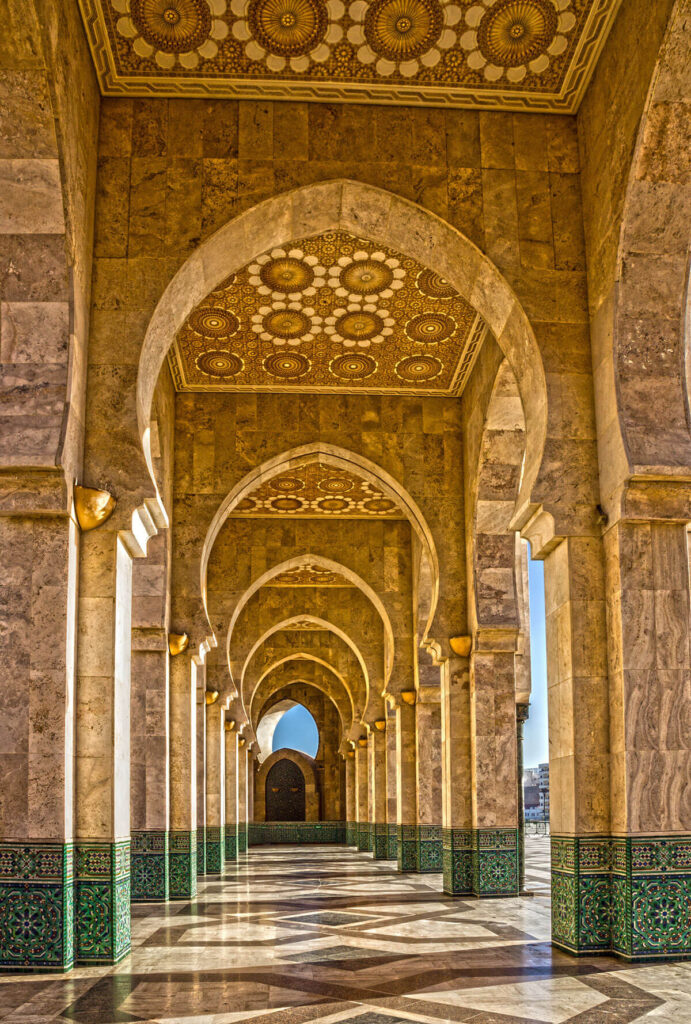 Ornate stone arches with intricate ceiling patterns and green-tiled bases form a corridor in a mosque, lit by sunlight streaming through the arches.