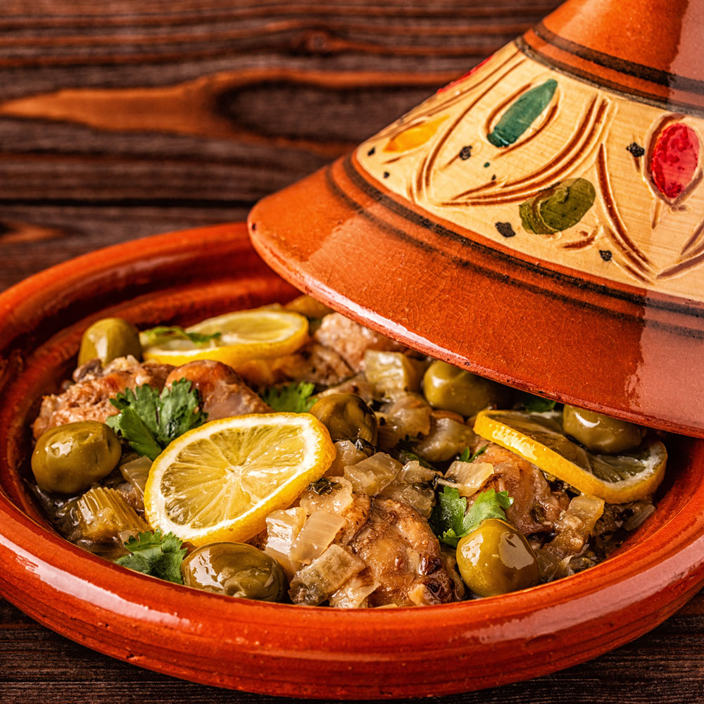 A Moroccan tagine with chicken, green olives, lemon slices, and herbs is served in a decorated clay pot with the lid partially open.