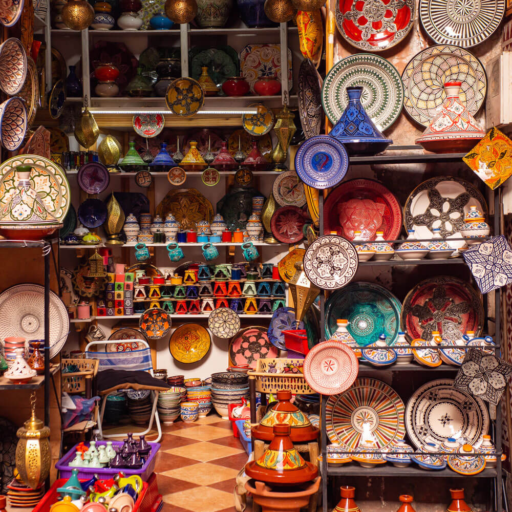 Shelves filled with colorful, patterned ceramic plates, bowls, and tagines in a market shop—perfect inspiration as you plan your luxury culinary or heritage trip to Morocco.