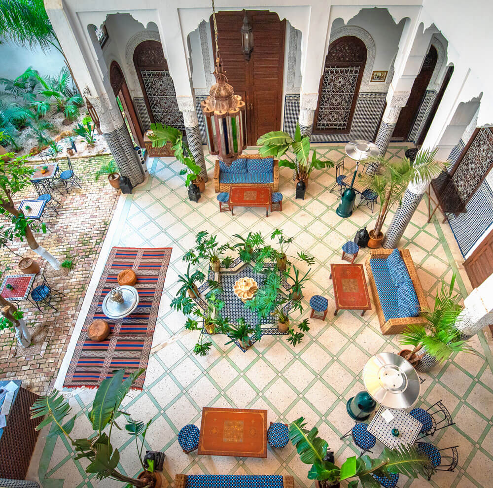 A courtyard with mosaic tile flooring, potted plants, blue cushioned seating, wooden tables, and decorative columns in a traditional Moroccan architectural style—perfect inspiration to plan your luxury culinary or heritage trip to Morocco.
