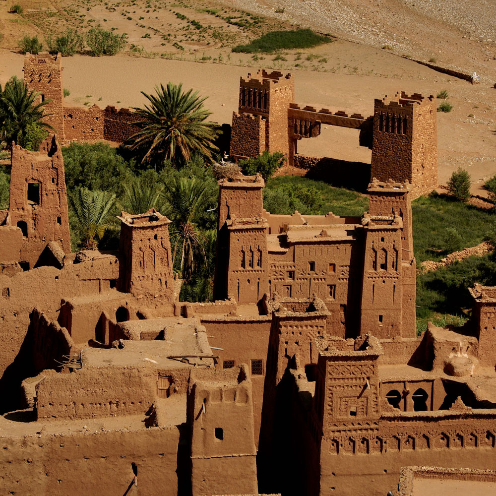 Plan your luxury culinary or heritage trip to Morocco and discover ancient earthen buildings with towers and geometric patterns, surrounded by palm trees and lush vegetation in a breathtaking desert landscape.