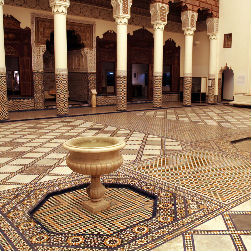 Ornate courtyard with geometric mosaic tiles, a central stone fountain, and columns supporting archways in a historic building—perfect inspiration as you plan your luxury culinary or heritage trip to Morocco.