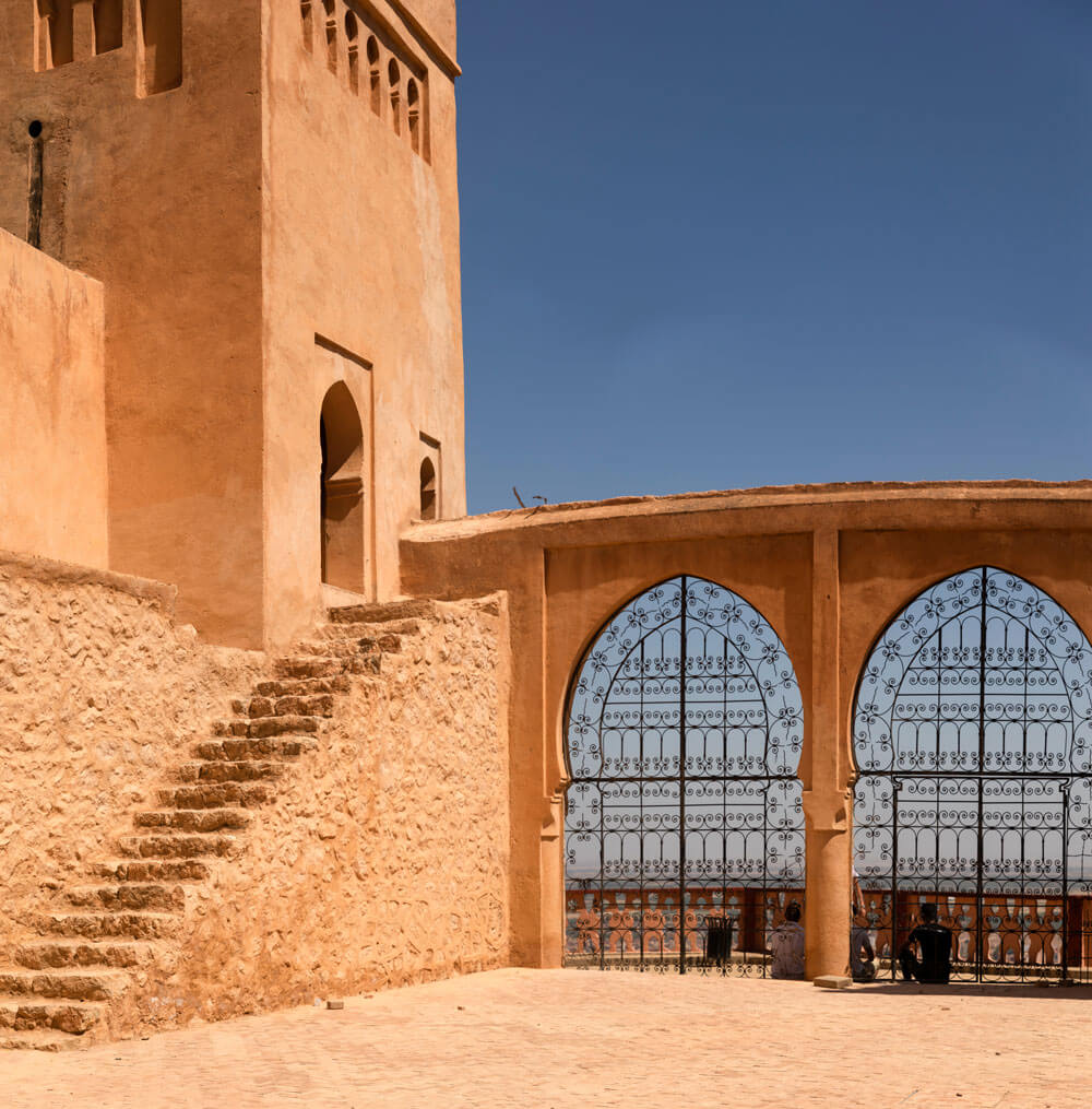 A stone stairway leads to an arched doorway beside ornate metal gates set in grand arches, beneath a clear blue sky—plan your luxury culinary or heritage trip to Morocco and experience stunning architecture like this.