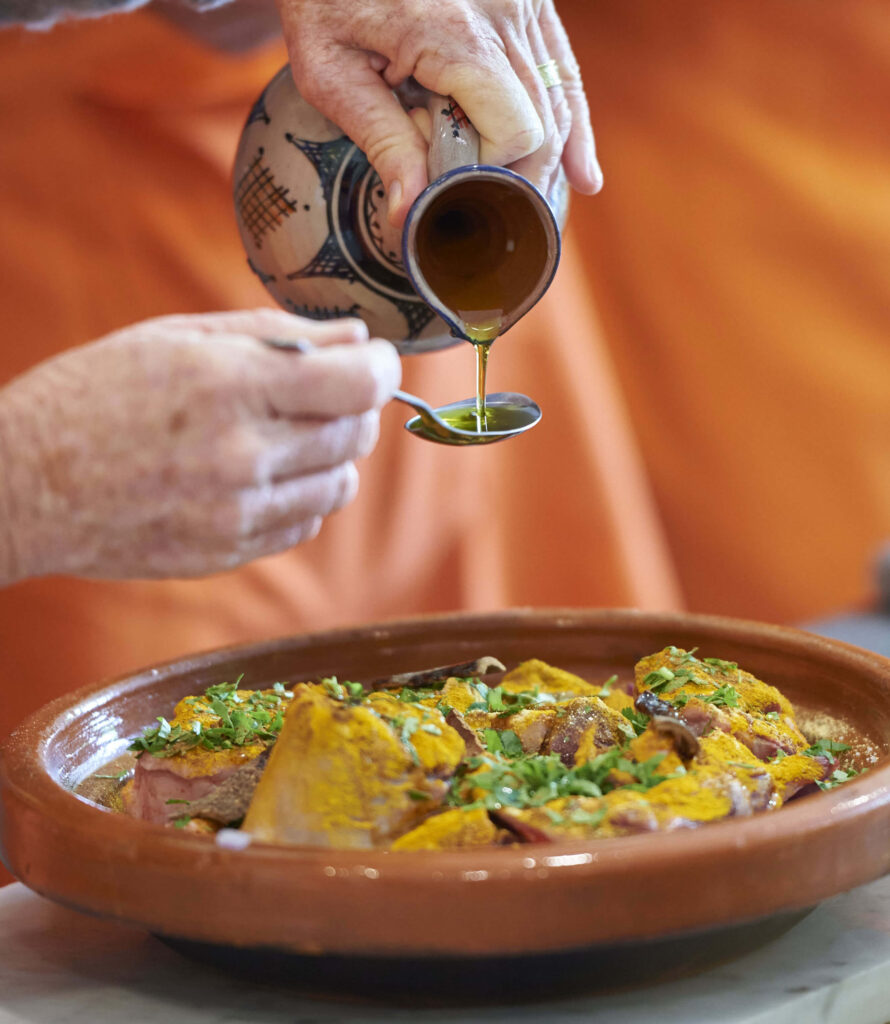 A person pours oil from a small jug into a spoon over marinated meat with herbs and spices in a brown ceramic bowl—a perfect scene for showcasing Morocco travel trade experiences.