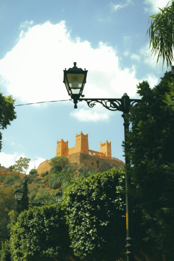 A stone castle with multiple towers sits atop a hill, framed by trees and street lamps under a partly cloudy sky.