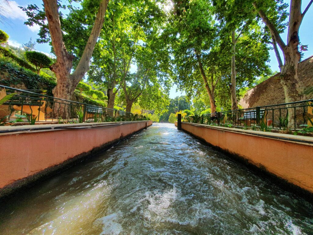 A narrow water canal flows between stone walls and railings, lined with tall leafy trees under a bright sky.