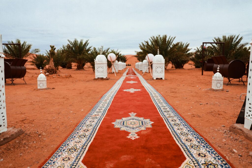 A patterned red carpet runs through a sandy desert with lanterns and palm trees lining both sides under an overcast sky.