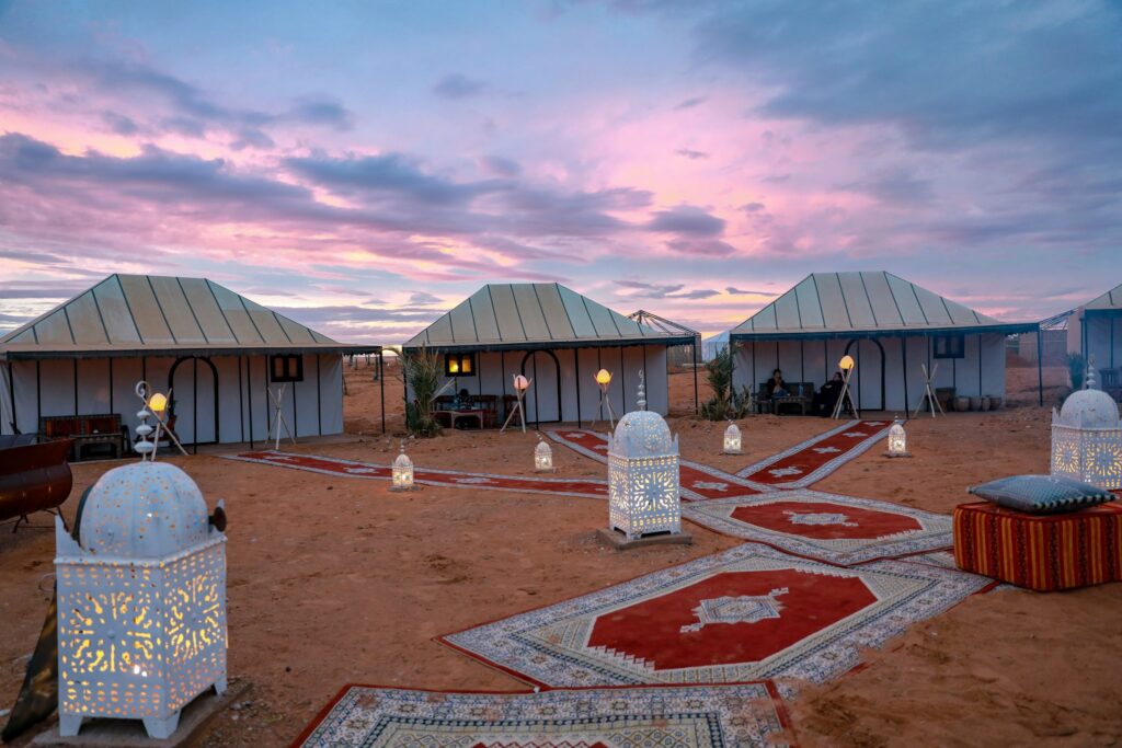 Four white tents are arranged on a sandy landscape at sunset, with patterned rugs and ornate lanterns placed on the ground.