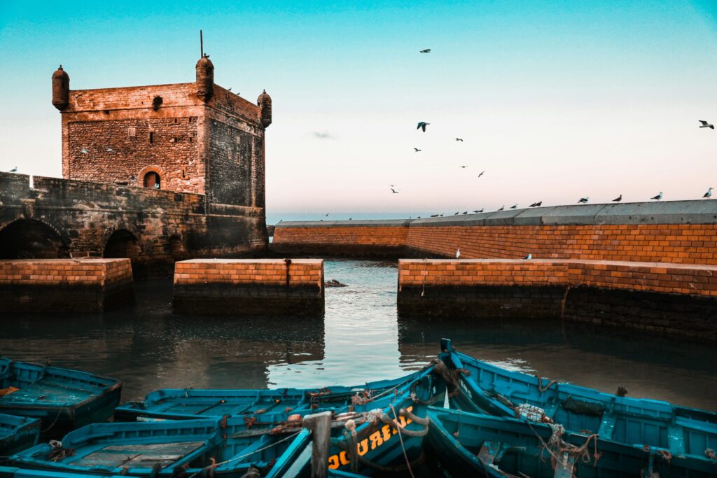 Stone fortress by a harbor with birds flying in the sky and several blue wooden boats docked in the foreground.