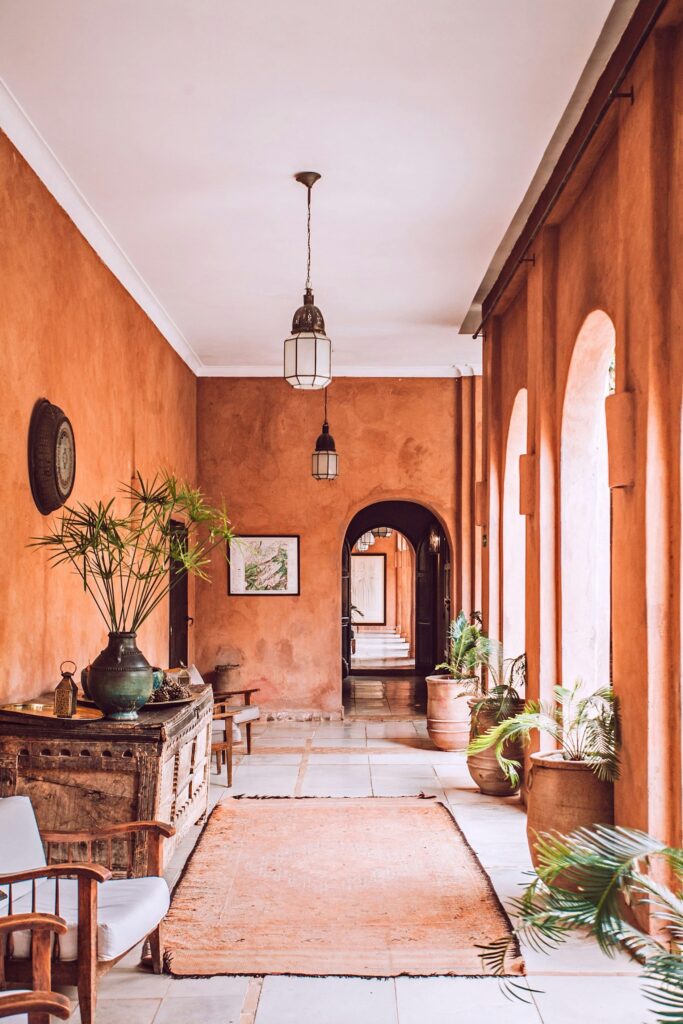 Long corridor with terracotta walls, arched windows, indoor plants, wooden furniture, a rug on the floor, and pendant lights hanging from the ceiling.