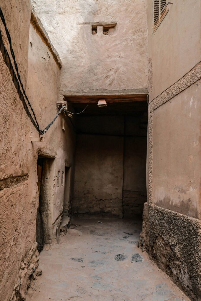 Narrow alleyway with textured, worn beige walls and a small overhang, showing exposed wires and weathered surfaces.