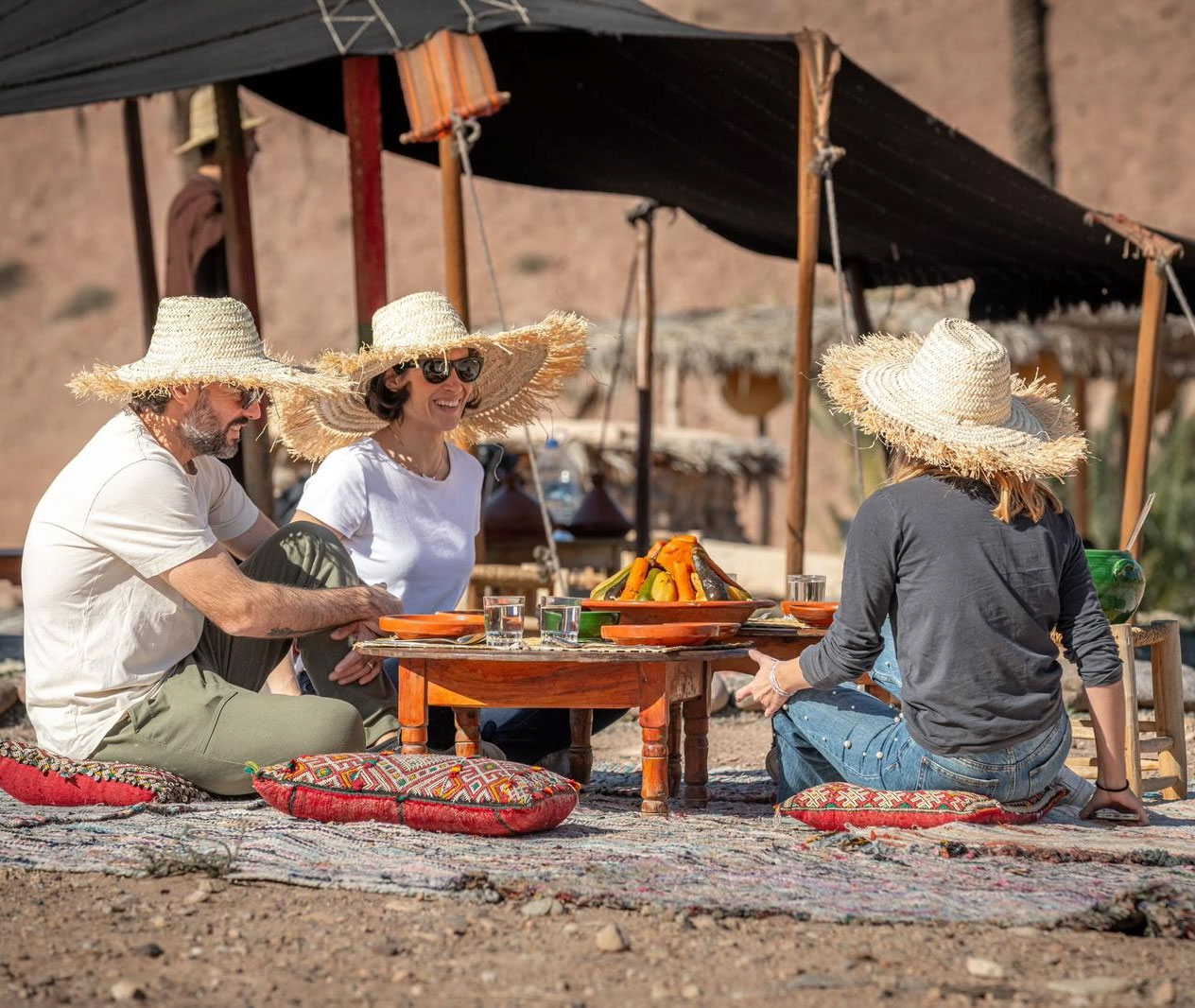 Three people wearing straw hats sit on cushions around a low table with food and drinks, outdoors on a patterned rug under a canopy in a desert setting.