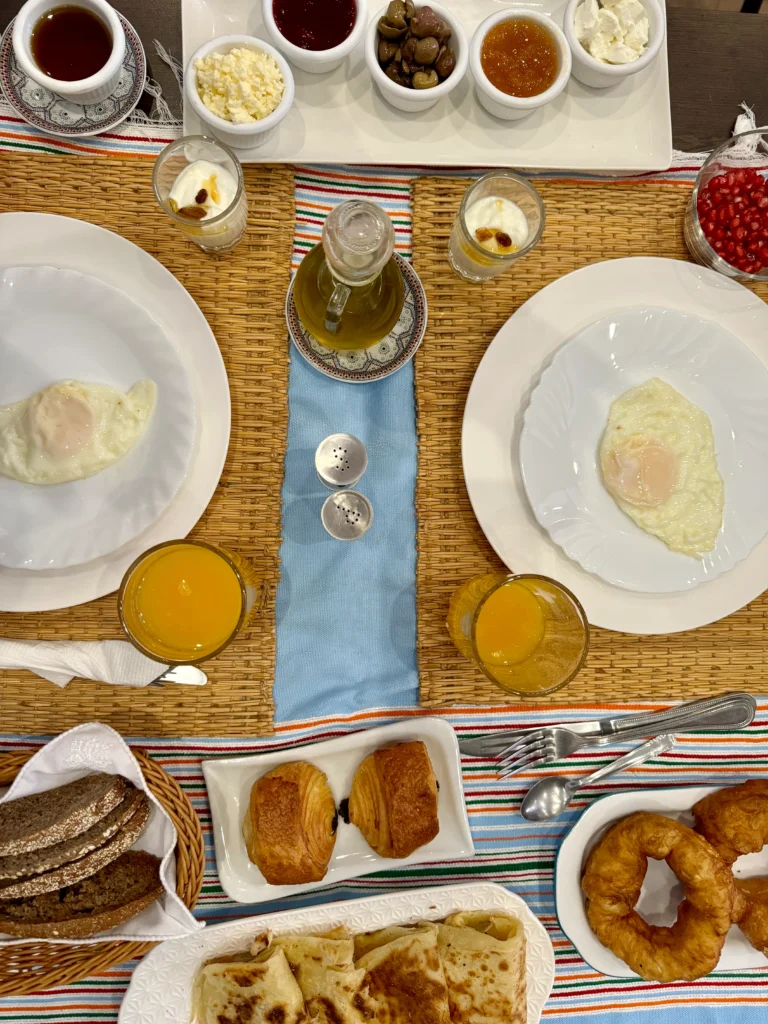 A breakfast table set for two with fried eggs, orange juice, bread, pastries, fried rings, flatbread, small dishes of cheese, jams, olives, yogurt, and pomegranate seeds.