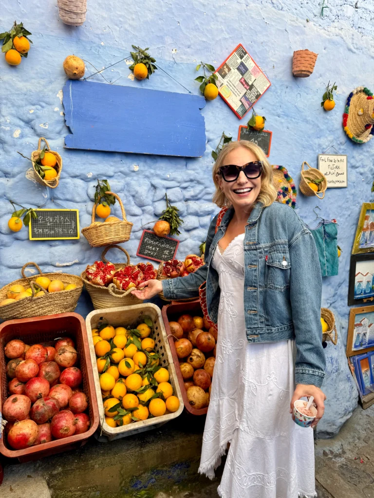 A woman in a white dress and denim jacket smiles while standing by baskets of pomegranates and oranges displayed against a blue wall.