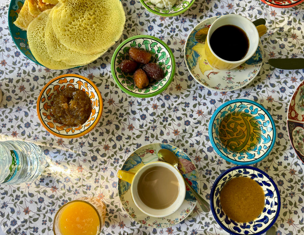 A table set with patterned dishes holding bread, dates, jam, drinks, and juice on a floral tablecloth. Two cups of coffee or tea are also present.