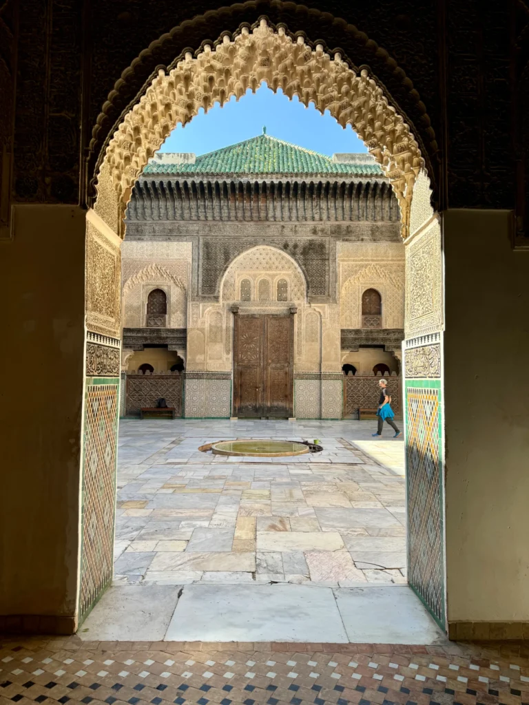 An arched doorway opens to a tiled courtyard with a central fountain, ornate walls, and a person walking past in front of a wooden door under a green-tiled roof.