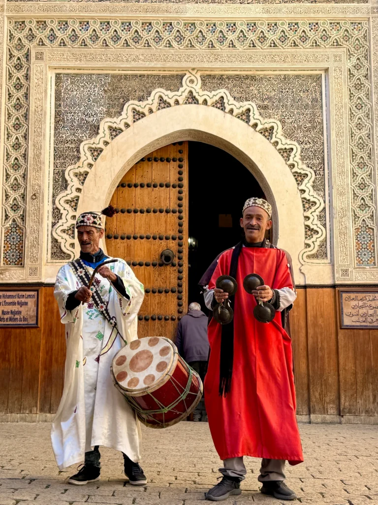 Two men in traditional attire play musical instruments in front of an ornate arched doorway with intricate tilework and wooden doors.