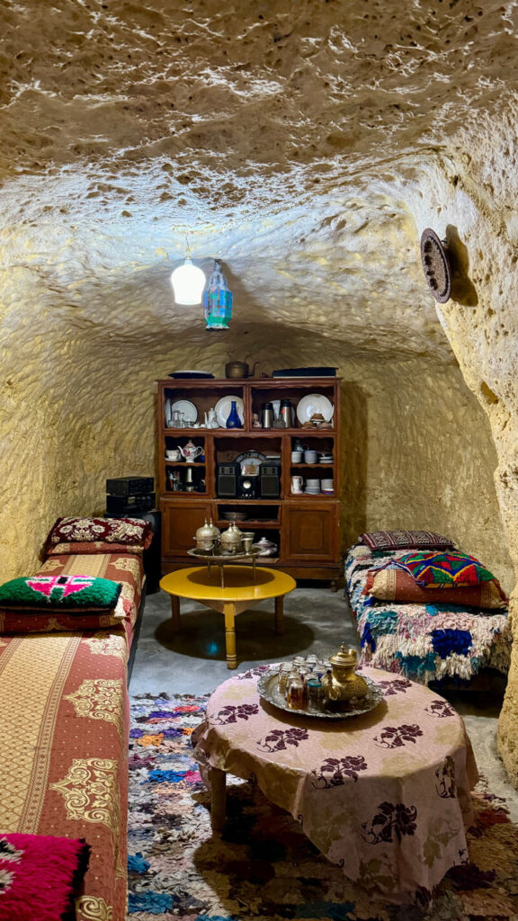 A cozy room carved into a cave, featuring two beds with colorful blankets, a wooden cabinet with dishes, and tables with tea sets under warm lighting.