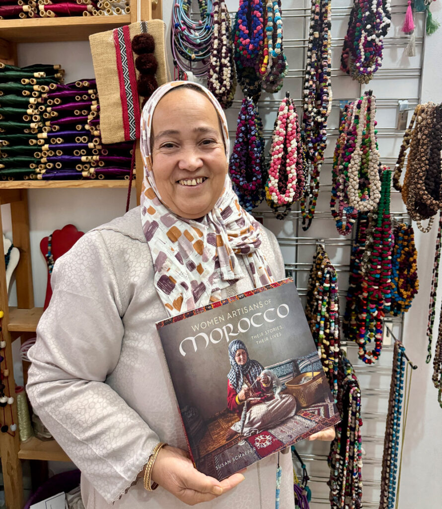A woman wearing a patterned hijab and coat stands in front of a display of colorful jewelry, holding a book titled “Women Artisans of Morocco.”.