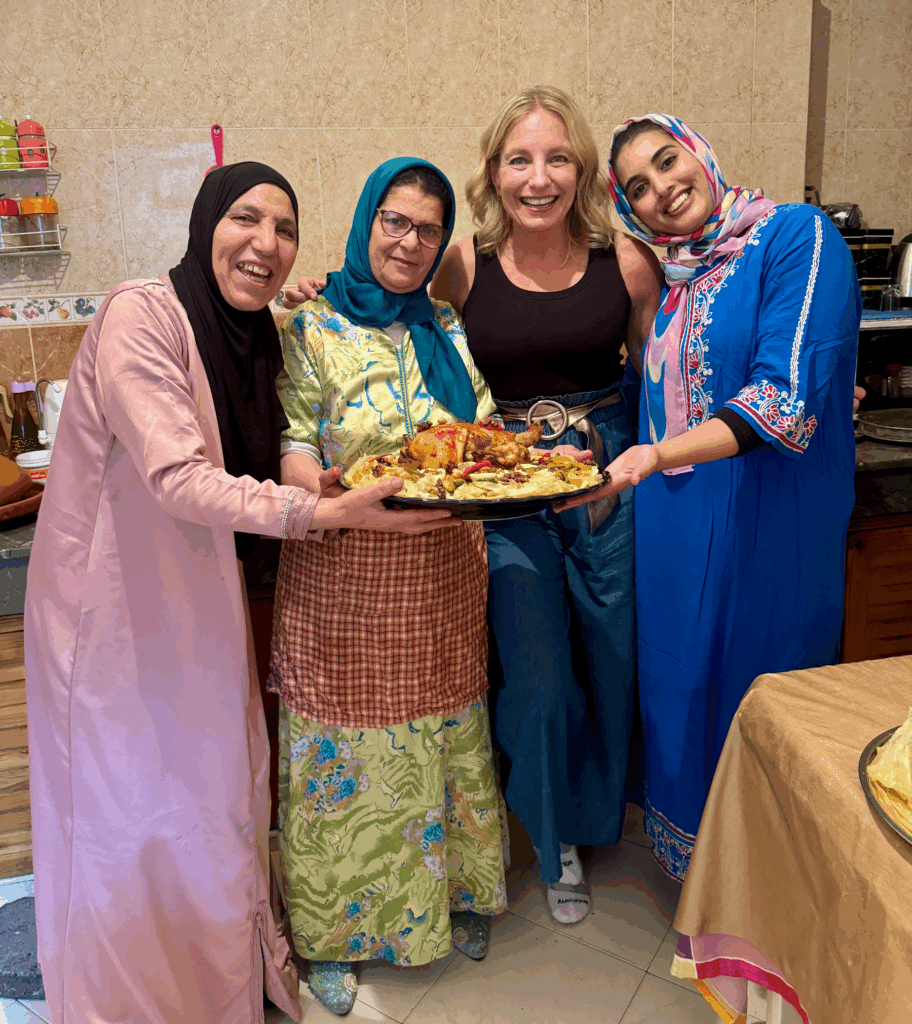 Four women stand together in a kitchen, smiling and holding a large platter of food, with tiled walls and kitchen items visible in the background.