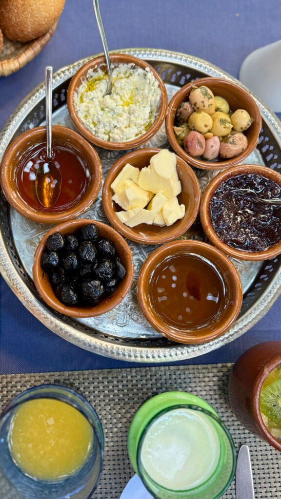 A round tray with six small bowls containing honey, cheese, olives, butter, olives, jam, and a spoon, set on a table with drinks nearby.