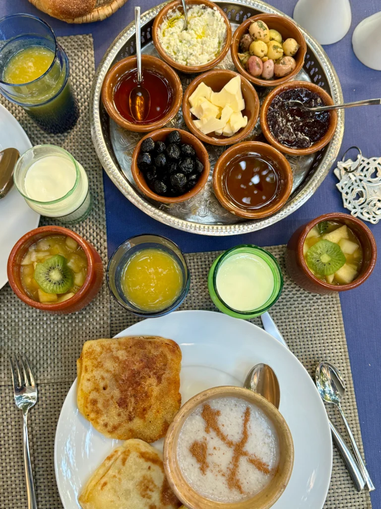 A table set with various small bowls of spreads, olives, butter, honey, and jam, alongside glasses of juice and yogurt, with bread and a plate of crepes and cereal in the foreground.