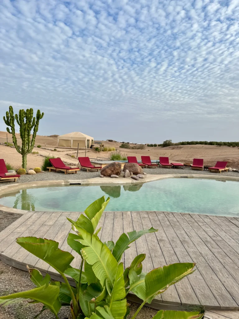 A round outdoor pool with red lounge chairs, a cactus, and desert landscape in the background under a cloudy sky. A tent structure is visible behind the pool.