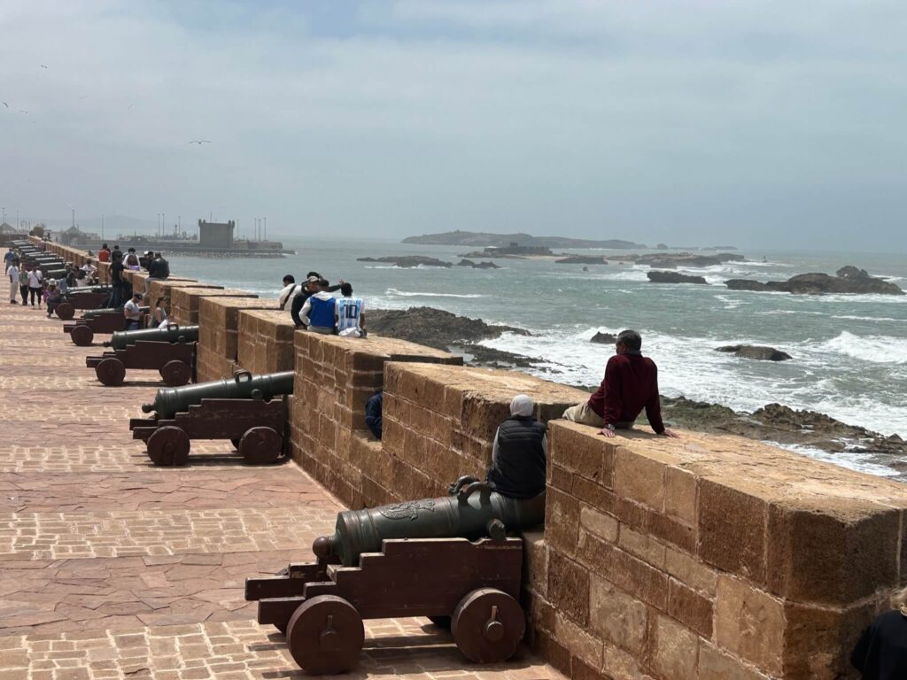 People sit and walk along a stone coastal fortification lined with old cannons, overlooking a rocky shoreline and the ocean under a cloudy sky—a perfect scene for exploring Morocco travel trade or forming B2B partnerships in an inspiring setting.