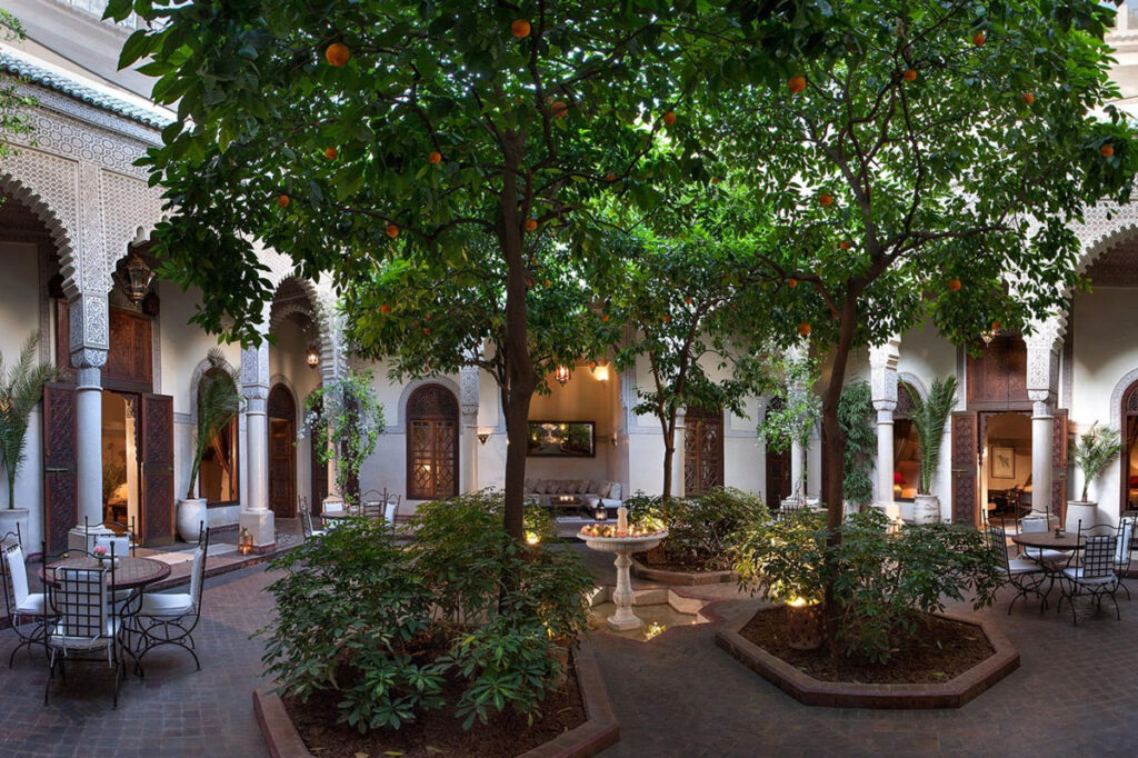 A courtyard with arched doorways, metal chairs and tables, lush greenery, orange trees, and a central stone fountain, surrounded by traditional architecture.
