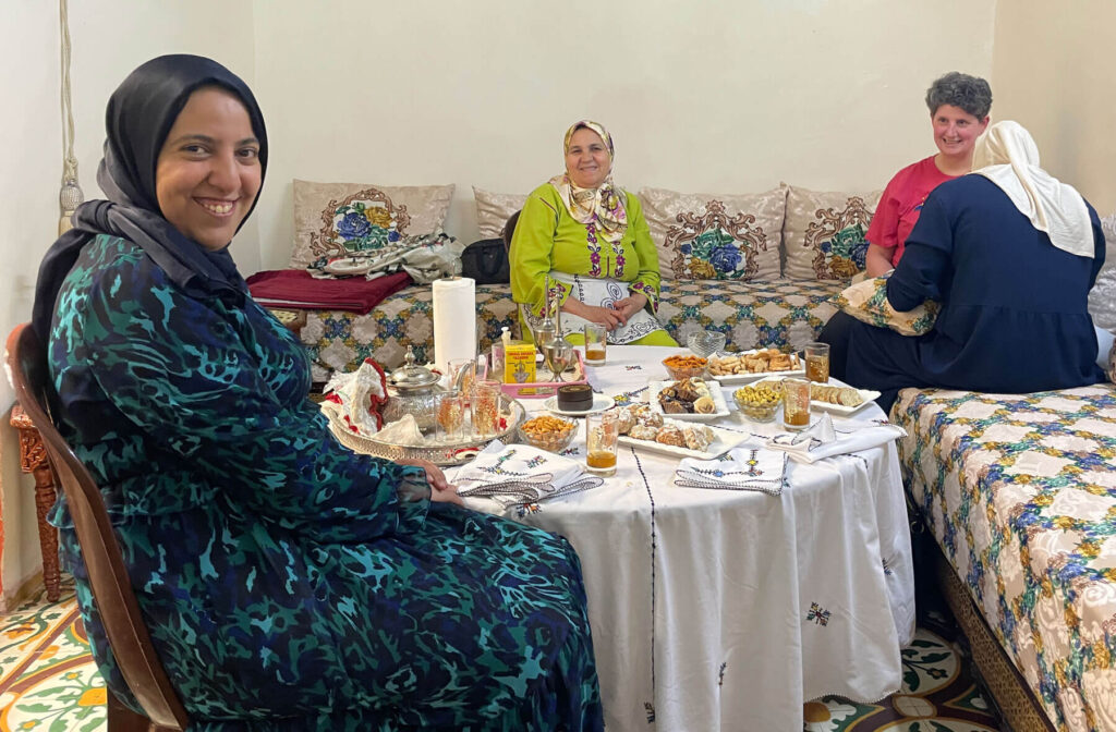 Four women sit around a low table covered with various dishes and drinks in a cozy, well-lit room with patterned cushions and bedding.
