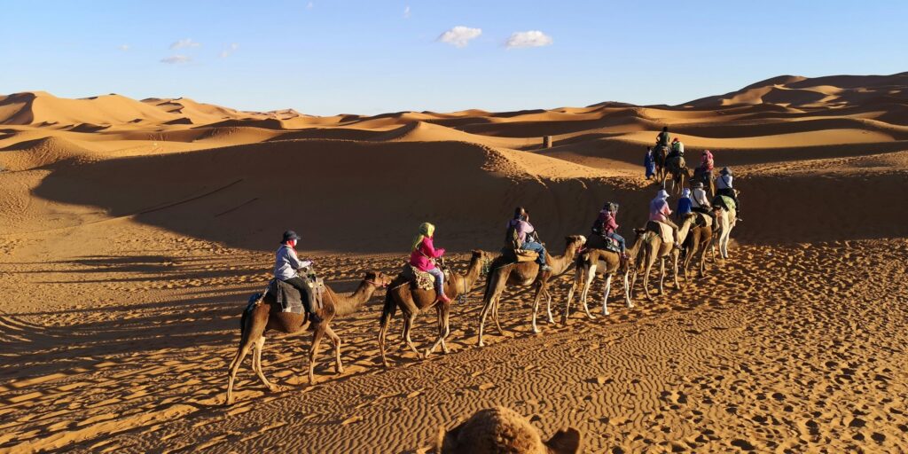 A group of people ride camels in single file across sand dunes in a desert under a clear blue sky.