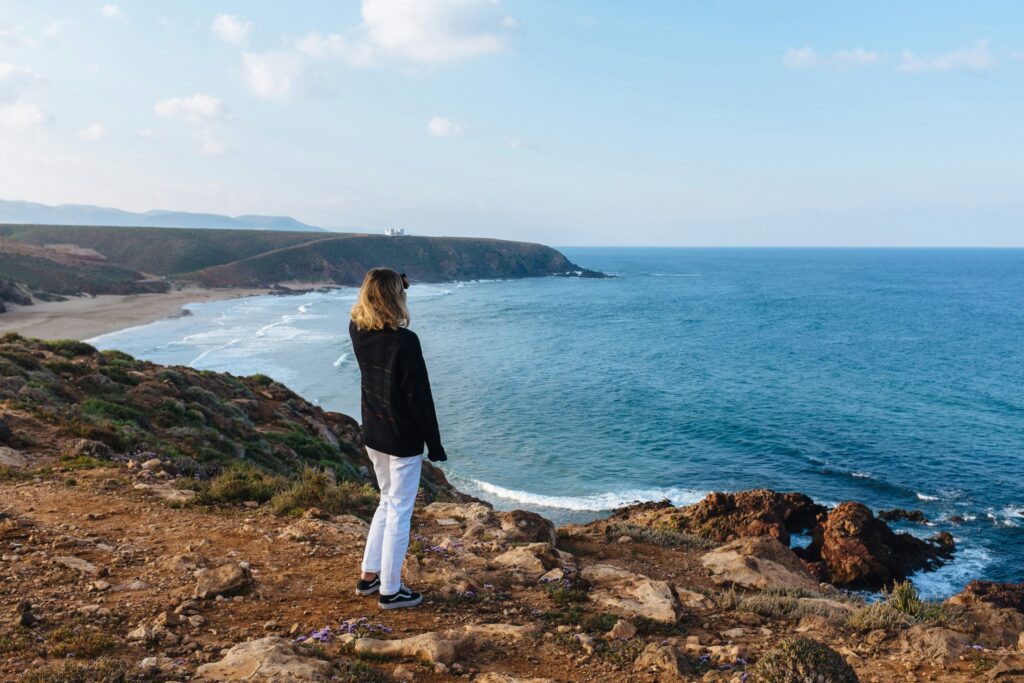 A person stands on a rocky cliff overlooking the ocean with waves meeting the shore and distant hills under a partly cloudy sky.
