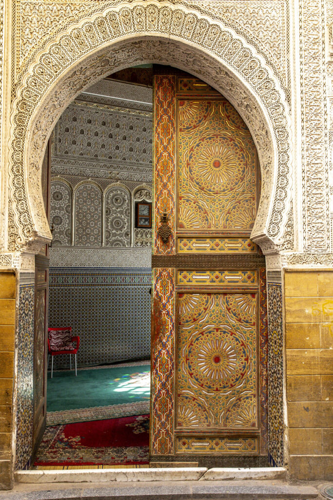 Ornate wooden door with intricate geometric designs opens into a room decorated with detailed mosaic tilework and patterned walls, revealing a red chair and green carpet inside.