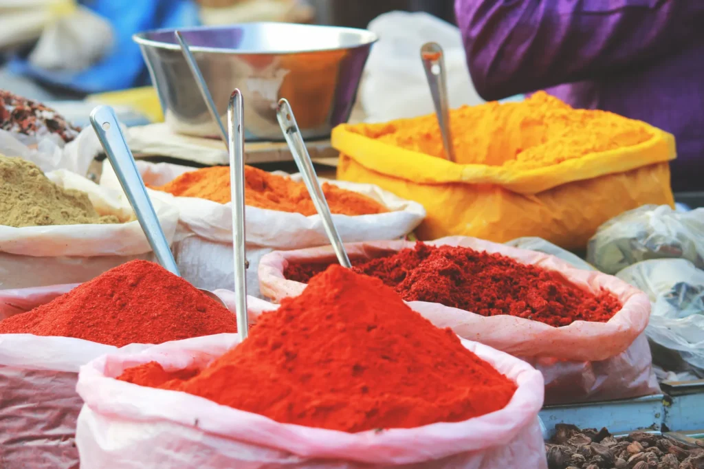 Bags of colorful powdered spices with metal scoops are displayed at an outdoor market stall.