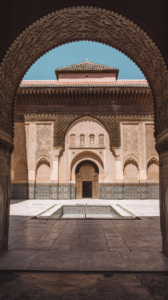 View through an ornate archway of a courtyard with patterned tile walls, a central rectangular fountain, and a wooden door in a historic building under a clear sky—perfect inspiration to plan your luxury culinary or heritage trip to Morocco.